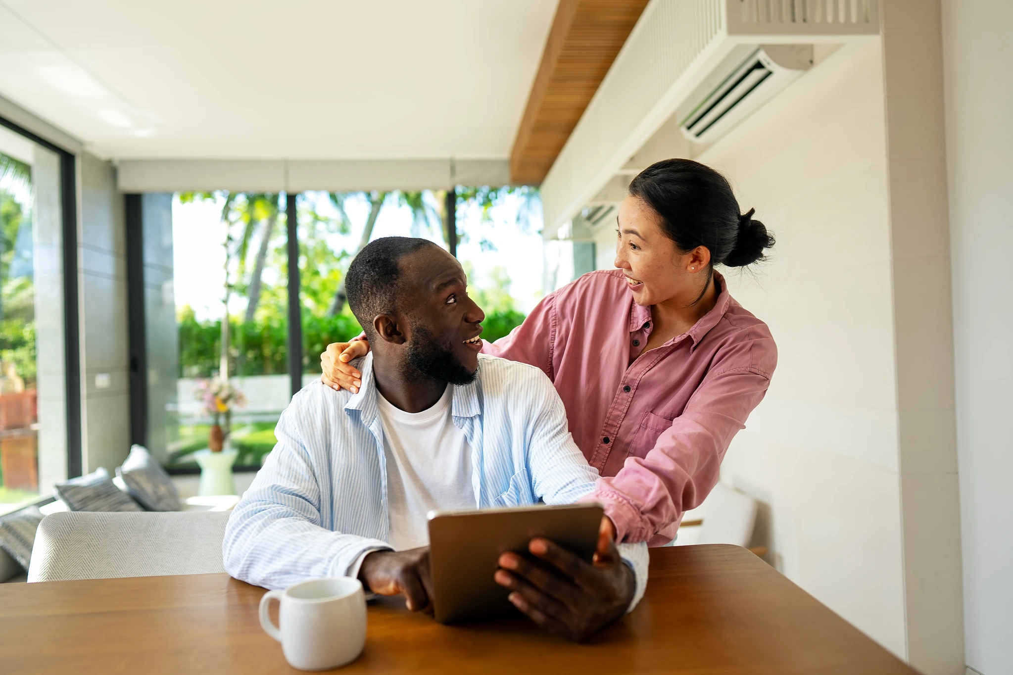 A man and woman smile at each other in a light-filled home. The man sits at a wooden table with a digital tablet in his hands and a white mug next to him. The woman has her arms around his shoulders. Both are wearing casual shirts. Large windows offer a view to trees outside. The photo evokes feelings of connection and joy, and could be used for projects about relationships, home life, technology, and togetherness.